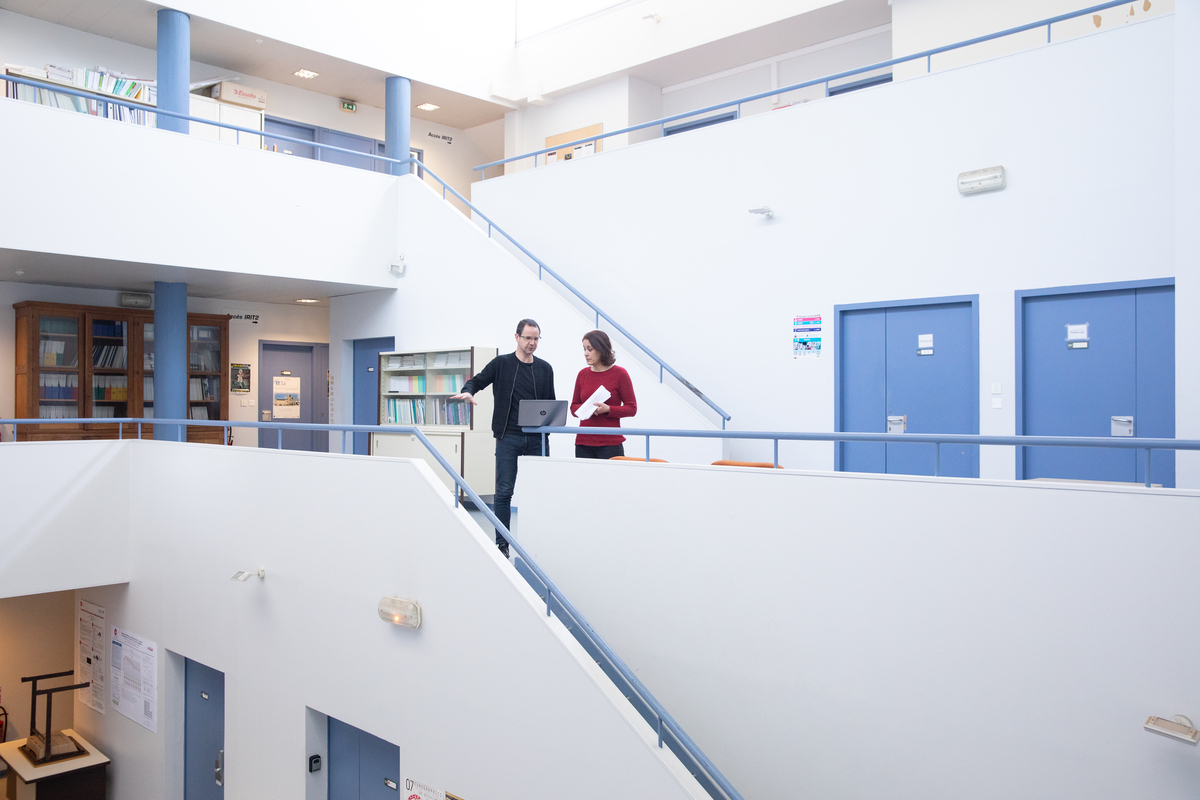 Un chercheur et une chercheuse au milieu des couloirs et escaliers d'un b&acirc;timent